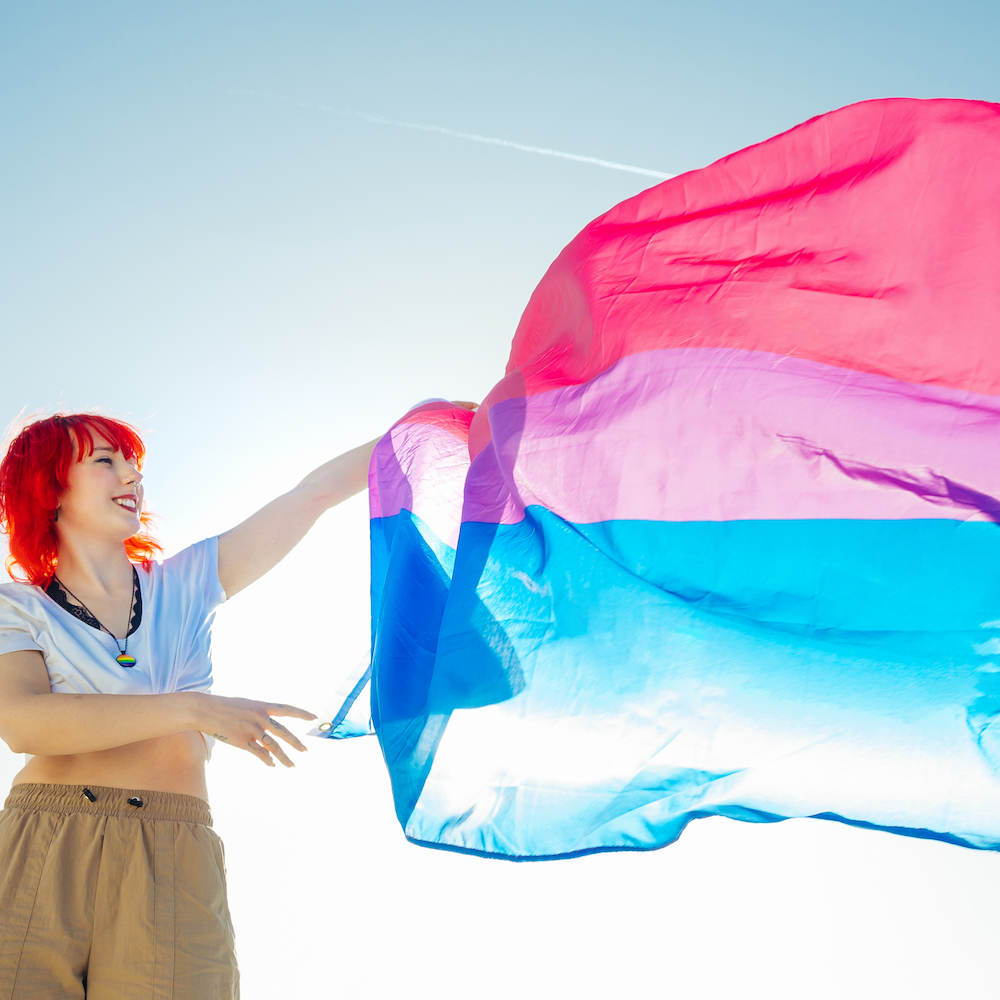 person with red hair holds a bi flag in the wind against a blue sky