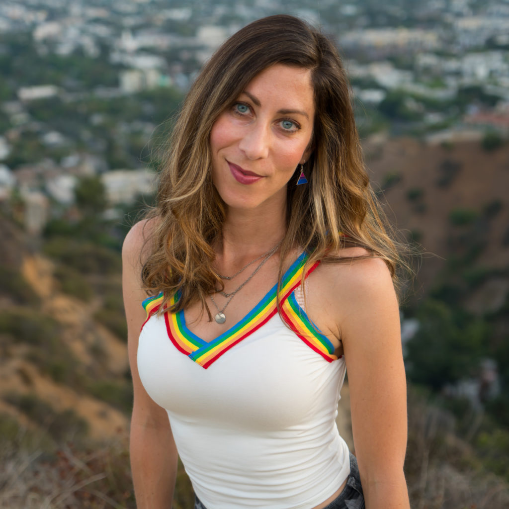 White person with shoulder-length brown and blond hair wearing a white and rainbow tank top posing on top of a hill with a city in the background
