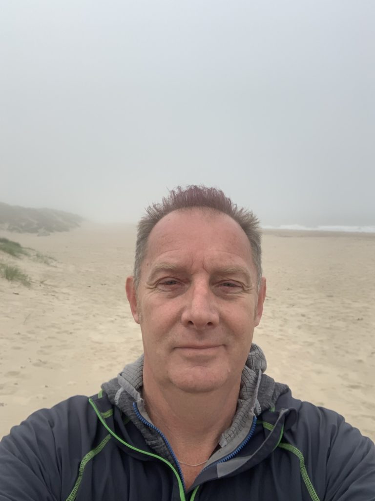 White man with short light hair taking a selfie with beach in background