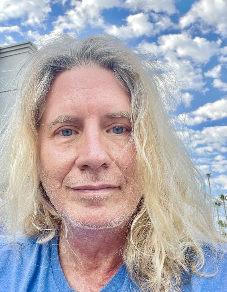 White person with light colored longer hair taking a selfie with blue sky and fluffy clouds behind
